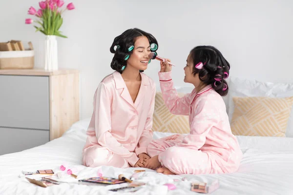 Menina adolescente chinesa alegre com encrespadores no cabelo aplica cosméticos na cara da mulher milenar no pijama rosa — Fotografia de Stock