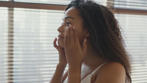 Soins de la peau. Jeune femme afro-américaine appliquant de la crème nourrissante sur ses joues, souriant à elle-même à la salle de bain, zoom avant — Video
