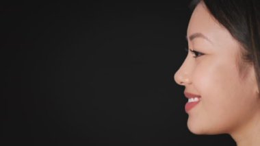 Kusursuz ciltli Asyalı genç bir kadının profil portresi gülümsüyor, siyah arkaplanda boş boşluğa bakıyor.