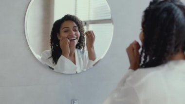 Ağız temizliği kavramı. Genç Afro-Amerikalı kadın bornoz giyiyor, dişlerini temizliyor, aynaya bakıyor.