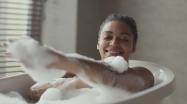 Genç mutlu Afrikalı Amerikalı kadın vücudunu süngerle yıkıyor köpüklü bir banyo yapıyor ve gülümsüyor.