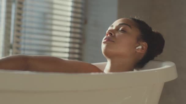 Ung afrikansk amerikansk dam njuter av koppla av i varmt bad med slutna ögon, lyssna på musik, selektivt fokus — Stockvideo