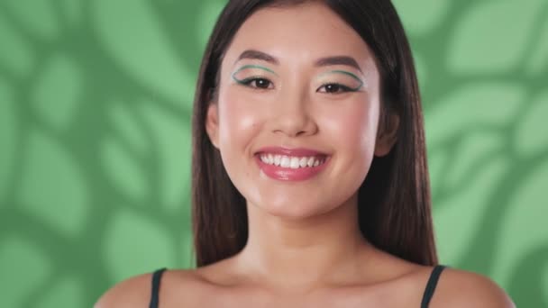 Kreatywny makijaż. Młoda piękna azjatycka kobieta z zielonymi strzałkami na oczach uśmiechnięta do kamery, zielone tło studio — Wideo stockowe