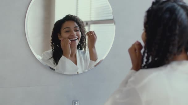 Поняття гігієни ур. Молода афроамериканка, одягнена в халат, чистить зуби, дивлячись на дзеркало у ванній кімнаті. — стокове відео