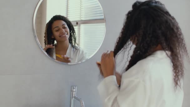 Концепція догляду за волоссям. Масштаб в портреті молодої безтурботної афроамериканки, що розчісує її кучеряве волосся спеціальним пензлем — стокове відео