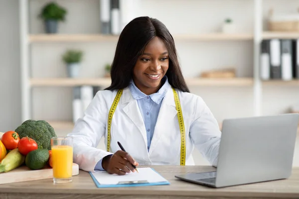 온라인에서 영양학 자에게 상담을 받았습니다. 친절 한 흑인 여성 영양사가 인터넷으로 노트북을 사용하고 필기를 하는 모습 — 스톡 사진