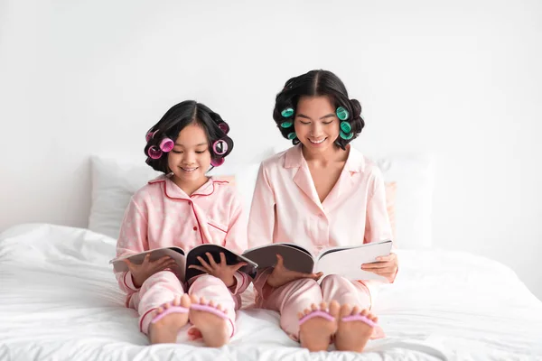 Lächelnde junge asiatische Frau und Teenie-Mädchen mit Lockenwicklern beobachten Magazin und Pediküre auf dem Bett — Stockfoto