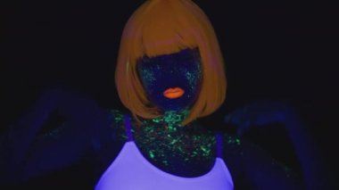 Gece kulübü tarzı. Parlak neon makyajlı, turuncu elbiseli ve ultraviyole ışıkta dans eden beyaz tişörtlü genç bir kadın.