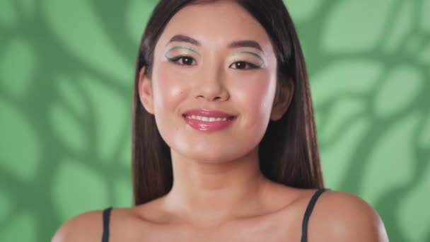 Junge verspielte asiatische Dame mit kreativem Make-up, die vor laufender Kamera einen Schlag abgibt und lächelnd über grünen Hintergrund flirtet — Stockvideo