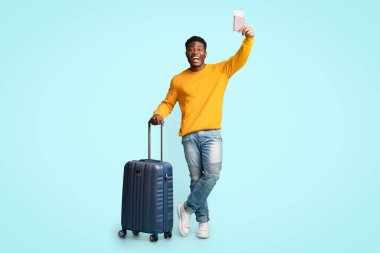Bavullu neşeli Afro-Amerikalı adam uçuş biletlerini gösteriyor.