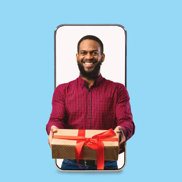 Glimlachende duizendjarige Afrikaans-Amerikaanse man geeft geschenk in doos feliciteren met vakantie op leeg scherm van enorme telefoon — Stockfoto