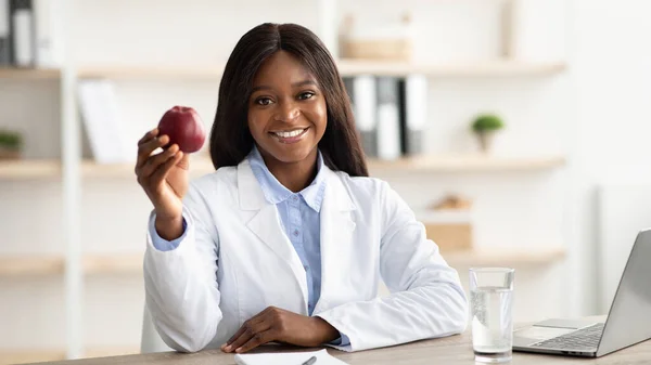 Piuttosto afro-americana nutrizionista femminile seduta davanti al computer portatile e con in mano mela rossa, che lavora in clinica — Foto Stock