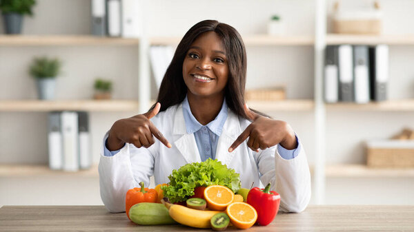 Portrait of happy african american dietitian pointing at fresh fruits and vegetables, smiling at camera at clinic
