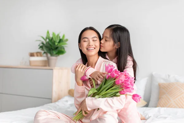 Улыбающаяся счастливая азиатская девочка-подросток целуется и обнимает тысячелетнюю женщину с букетом тюльпанов в спальне — стоковое фото