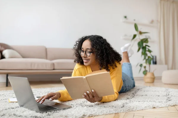 Joven mujer negra leyendo libro cerca de la computadora portátil, preparándose para el examen o trabajando en el proyecto, acostado en el suelo en casa —  Fotos de Stock