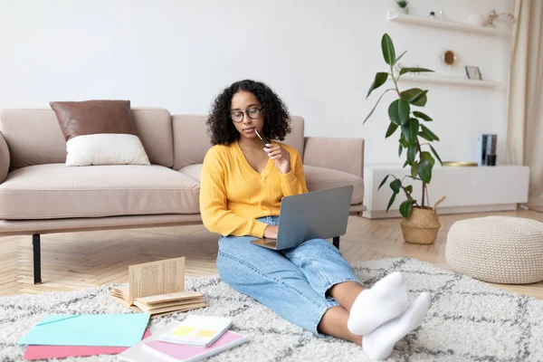 Jonge zwarte vrouw zitten op de vloer met studiemateriaal, het hebben van online klasse met tutor via laptop computer van thuis — Stockfoto