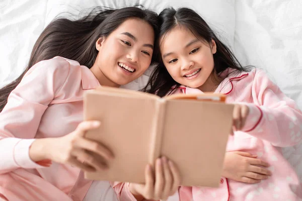 Sonriente asiática joven mujer y adolescente chica en pijama rosa acostado en la cama mira el libro en la cama en el dormitorio, vista superior —  Fotos de Stock