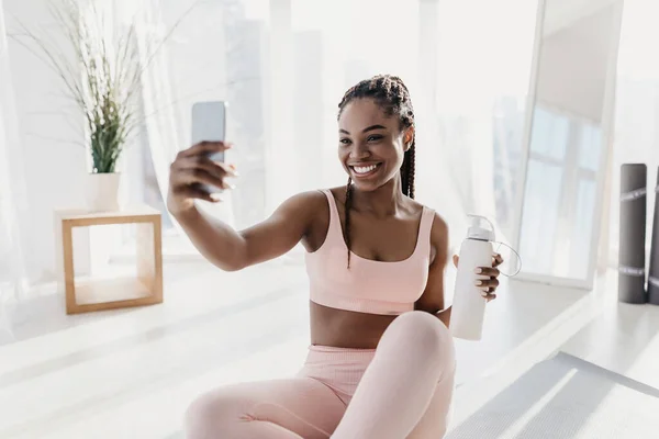 Encantadora jovem afro-americana com garrafa de água tomando selfie durante o treino em casa — Fotografia de Stock