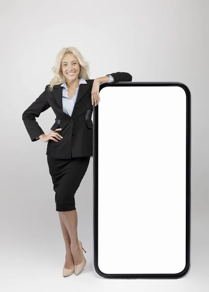 Selbstbewusste junge Geschäftsfrau lehnt an einem großen Smartphone mit weißem leeren Bildschirm und zeigt freien Platz für Werbe- oder App-Design — Stockfoto