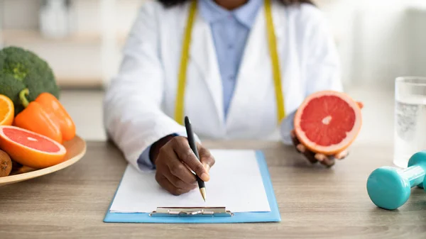 Dietóloga afroamericana sosteniendo toronja y escribiendo un plan de alimentación saludable o dieta para perder peso para el paciente —  Fotos de Stock