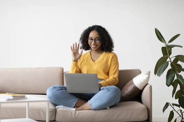 Comunicación en línea. Mujer negra en gafas sentada en el sofá en casa, saludando a la webcam, teniendo videollamada en casa —  Fotos de Stock