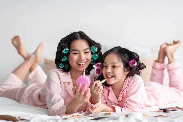 Allegro millenario signora giapponese con bigodini tenere specchio per teen girl, applicare il rossetto sulle labbra, sul letto con cosmetici — Foto Stock