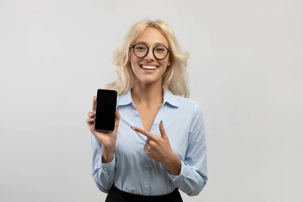 Бізнес-додаток. Щаслива бізнес-леді тримає смартфон і вказує палець на порожній екран, ліга сірий студійний фон — стокове фото