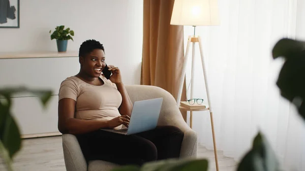 Ung positiv afrikansk amerikansk dam talar på mobiltelefon och arbetar på bärbar dator, konsultera kunder hemifrån — Stockfoto
