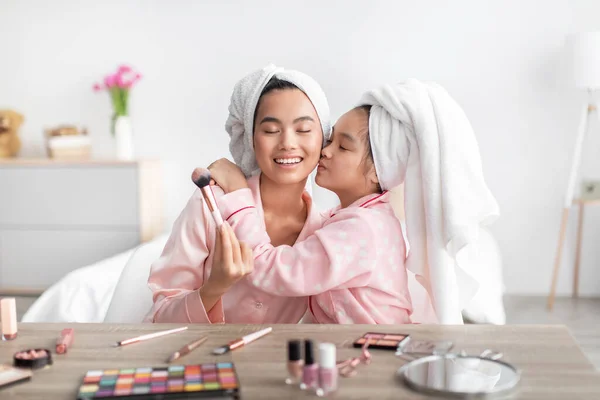 Puas Korea gadis remaja mencium wanita muda dalam piyama merah muda dan handuk di kepala di meja dengan kosmetik — Stok Foto