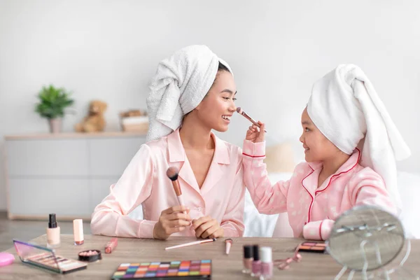 Gadis remaja cina yang ceria dan perempuan muda dengan piyama merah muda dan handuk di kepala melakukan makeup di kamar tidur — Stok Foto