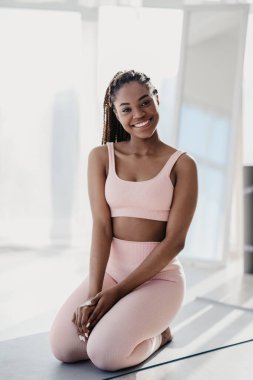 Gülümseyen Afrikalı Amerikalı kadın spor kıyafetleri giyiyor, spor paspasına oturuyor, antrenmanlara ara veriyor.