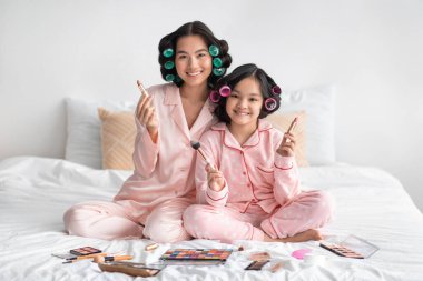 Mutlu milenyum Japon kadın ve bigudili ergen kız fırça tutarak yatakta kozmetikle otururlar.