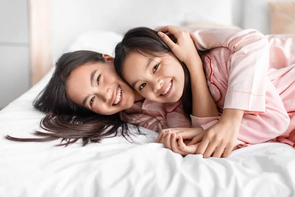Feliz feliz bastante lindo japonés joven mujer abrazo adolescente chica en rosa pijama mentira en la cama y divertirse en dormitorio —  Fotos de Stock