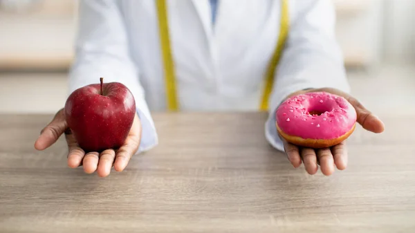 黒栄養士はドーナツとリンゴを保持し、新鮮で不健康な食べ物の間で選択を提供し、職場に座って — ストック写真