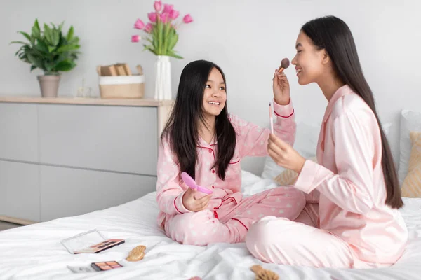 Allegro felice coreano giovane ragazza applicando polvere per viso donna millenaria, seduto sul letto con cosmetici — Foto Stock