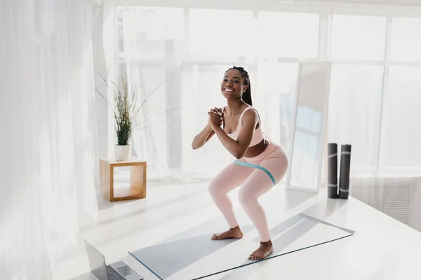 Longitud completa de ajuste joven mujer afroamericana en cuclillas con banda elástica en la estera, ejercicio en línea en casa, espacio de copia —  Fotos de Stock