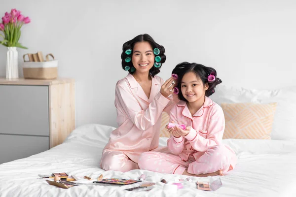 Улыбающаяся китаянка делает прическу с бигуди для молодых девушек, сидящих на кровати с косметикой в спальне — стоковое фото