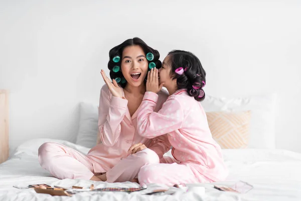 Schockierte junge Chinesin hört Klatsch und Tratsch von Teenie-Mädchen mit Lockenwicklern, sitzt mit Kosmetik im Schlafzimmer auf dem Bett — Stockfoto