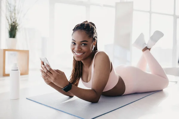 Черная женщина лежит на коврике для йоги, используя смартфон во время тренировки, носит наушники, слушает любимую музыку — стоковое фото
