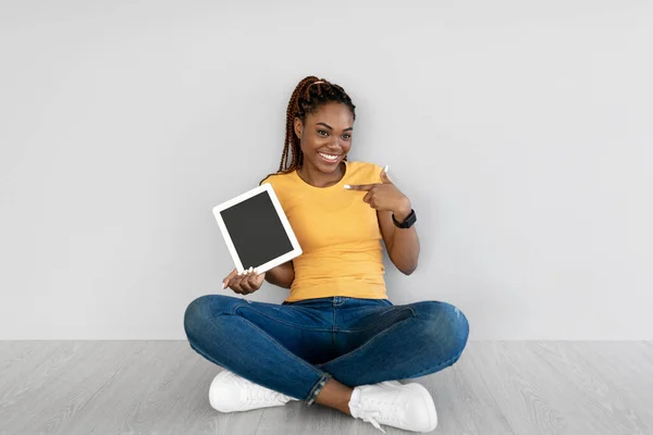 Fekete nő mutat tabletta üres képernyőn, ül a padlón szemben szürke stúdió fal, mockup honlap vagy alkalmazás — Stock Fotó