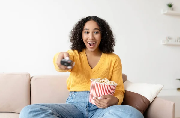 Portrét mladé černošky s dálkovým ovládáním sledování televize a jíst popcorn na gauči doma — Stock fotografie