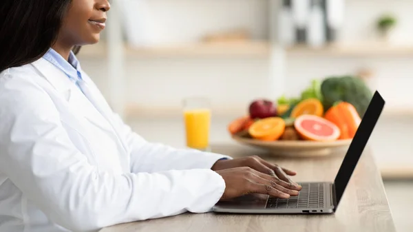 Μαύρη γυναίκα διαιτολόγος με φορητό υπολογιστή συμβουλευτική ασθενή σε απευθείας σύνδεση στην κλινική, κάθεται στο γραφείο, πλαϊνή άποψη, περικοπεί — Φωτογραφία Αρχείου