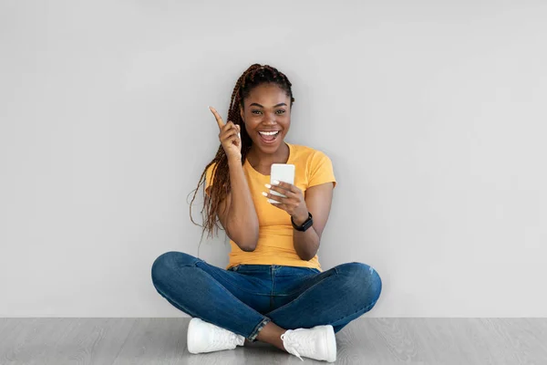 Emocionada joven afroamericana sentada en el suelo con smartphone, teniendo una idea creativa contra la pared gris del estudio — Foto de Stock