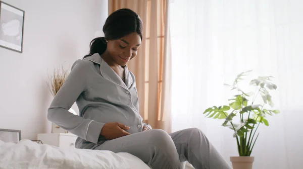 Jonge vreedzame Afro-Amerikaanse zwangere vrouw strelen haar buik en glimlachen, zitten op bed in de ochtend, lege ruimte — Stockfoto