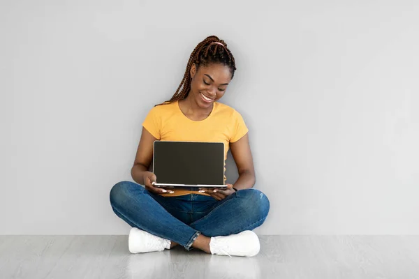 Lovely young black woman sitting on floor, showing laptop with empty screen near grey studio wall, mockup for website — Stock Photo, Image