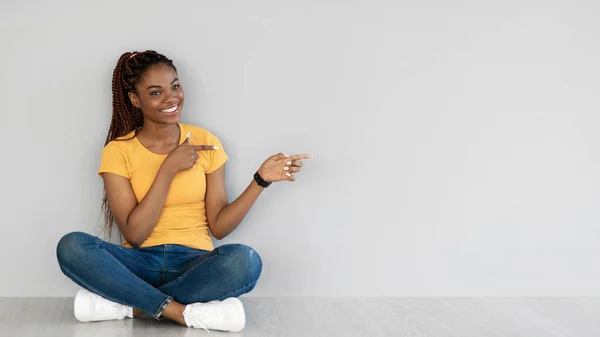 Veselá mladá černoška sedící se zkříženýma nohama na podlaze, ukazující stranou na volné místo u šedé zdi, prapor — Stock fotografie