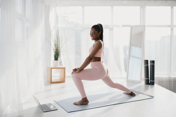 Hermosa joven mujer negra haciendo ejercicio delante de la computadora portátil en casa, viendo video tutorial, disfrutando de la formación doméstica —  Fotos de Stock