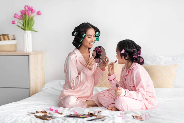 Felice giovane mamma cinese con bigodini tenere specchio per figlia adolescente, bambino applicare il rossetto sulle labbra sedersi sul letto — Foto Stock