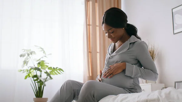Jovem feliz afro-americano em pijama sentado na cama e abraçando a barriga, desfrutando do tempo de gravidez em casa, espaço livre — Fotografia de Stock