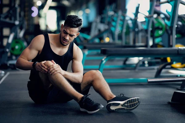 Trauma desportivo. Jovem atleta árabe masculino tocando seu joelho ferido no ginásio — Fotografia de Stock